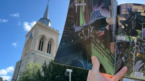 A hand holds open a Batman comic book outdoors. The comic shows dark, moody illustrations of Batman perched at a church in a city at night. In reality there is church tower with arched windows behind the open comic book.
