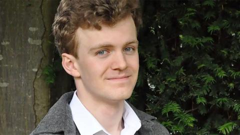 Sam Carling, who has brown hair, standing outside wearing a white shirt and a grey jacket