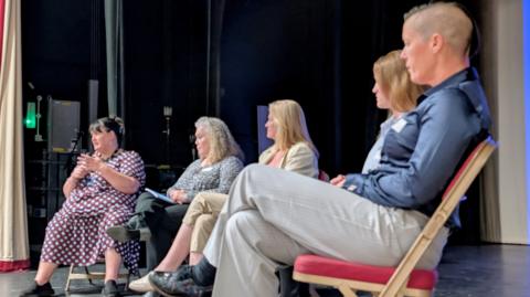 Picture of a panel in discussion on a stage, taken from the side. Four people sit on chairs in a semi-circle. One at the end is speaking to the audience.