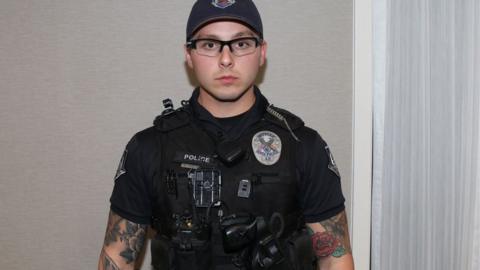 Pahilip Brailsford, dressed in police gear, wearing a cap and glasses. His arms are heavily tattooed and he is clean-shaven