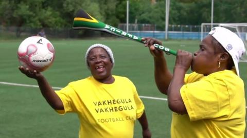 Two older women in yellow t-shirts on a football pitch, one holding a ball, the other a vuvuzela