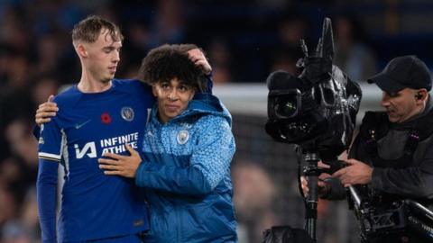 Cole Palmer of Chelsea embraces Rico Lewis of Manchester City filmed by a Sky Sports Steadicam cameraman after the Premier League match between Chelsea FC and Manchester City. Palmer is on the left wearing a blue Chelsea football shirt. Lewis is on his right wearing a light and dark blue Man City jacket. The cameraman is to their right holding a big black camera pointed at them from the side and he is dressed in black with a black cap on.