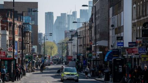 Wide shot of people walking down a street in Dalston in Hackney as a police car drives down the road