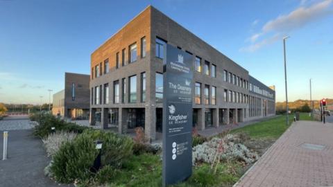 The Deanery CE Academy. It is a modern brick building with lots of windows surrounded by planted greenery