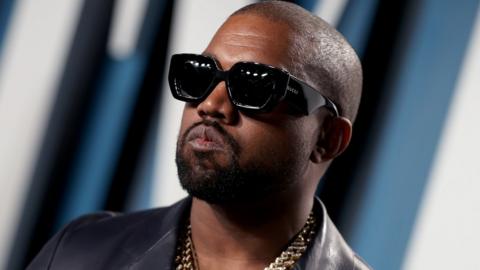 A headshot of Kanye West who is looking into the distance past the camera. he is wearing big black sunglasses and has a gold chain around his neck