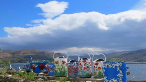 A large white cloud sits in a bright blue sky above mountains and a lake, with painted watering cans the the foreground