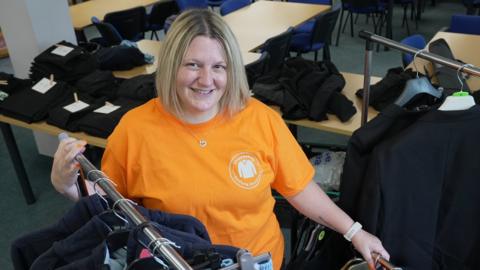 Woman in orange t-shirt looks up at camera surrounded by rails of school uniform.