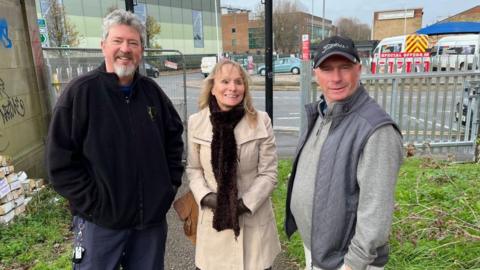 Two men either side of a woman. One man is wearing a cap. They are standing by the entrance to the footpath