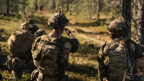 British troops wear camouflage army uniforms as they walk through a forest
