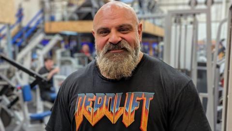 Laurence Shahlaei in a gym, wearing a black tshirt with the word Deadlift on it.