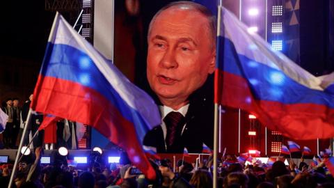 Vladimir Putin on a large screen as a crowd waving Russian flags watch on