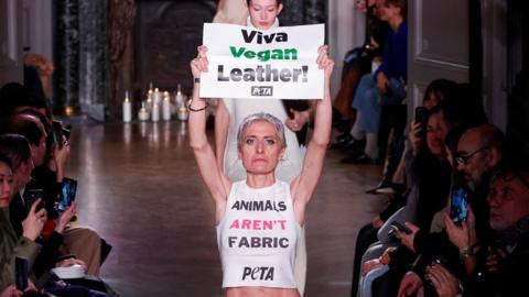 A Peta activist wearing a t-shirt that says "animals aren't fabric" and holding a sign that reads "viva vegan leather" blocks the runway at the Paris Fashion Week