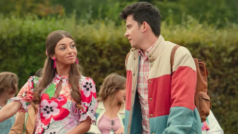 Mimi Keene as Ruby and Asa Butterfield as Otis in Sex Education. Ruby and Otis are speaking to each other outside as they wheel their bikes. Ruby, a young woman with long light brown hair, wears a short floral dress and large pink earrings. Otis, a young man with short dark hair, wears orange cord trousers, a pink plaid shirt and a blue, cream and red striped puffer jacket.