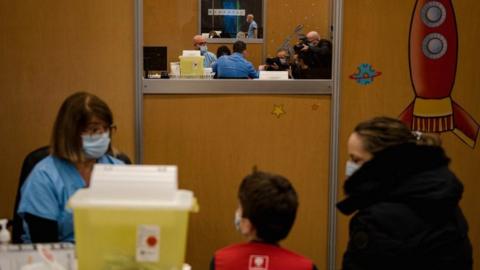 A child sits with his mother as he waits for his dose of the Pfizer-BioNTech vaccine for children as photographers photograph another child receiving the vaccine in November 2021 in Montreal, Quebec