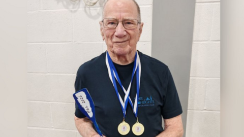 Peter Quinney, wearing two gold medals from his recent trampolining competition. He wears glasses and is smiling at the camera, wearing a navy t shirt. Behind him is a white brick wall.