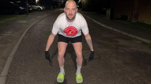 Daniel Fairbrother wearing bright green slider shoes on his feet, with white socks, crouching down, to show them, in a street, wearing a T-shirt, black shorts and black gloves. It is dark and you can just see cars and houses. 