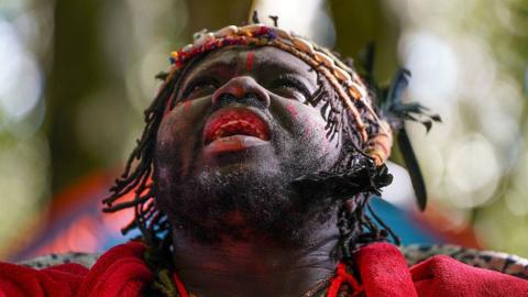 A man in a red top, with braided hair and a headpiece