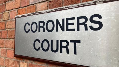 A silver plaque on the outside of the court. The words 'coroners court' are written in bold capital letters in black. The sign is on a brick wall. 