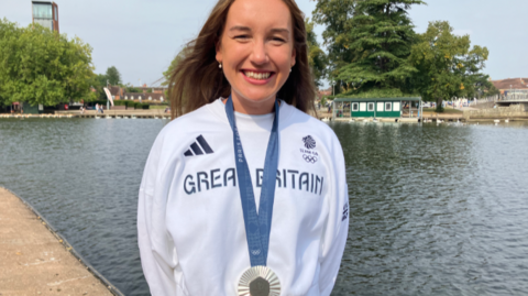 Esme is wearing a white Team GB top with her silver medal over the top and is standing by water in Stratford-upon-Avon