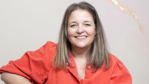 A woman with straight blonde hair and an dark orange shirt is smiling at the camera against a plain background