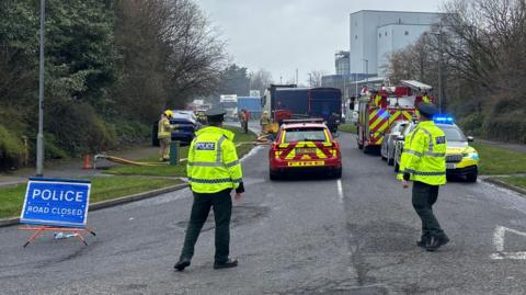 Two police officers standing at a junction of a road. There is a blue police sign to the left of the image and there is a fire car, a police car and a fire engine in front of them.