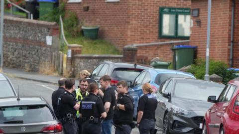 About half a dozen police officers stood in a residential street lined either side with cars