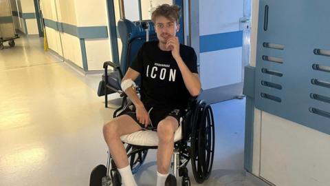 A man sits in a wheelchair in the hallway at Nicosia General Hospital, Cyprus. He has a black T-shirt on, black shorts and is wearing white socks. The corridor has white walls with light blue fittings. 