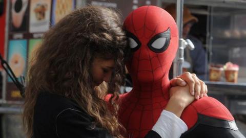 Tom Holland as Spider-Man holds Zendaya, who plays MJ, on set. Holland is dressed in a ful Spider-Man suit against a New York backdrop and Zendaya wears a black top and has long brown hair 