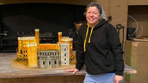 Michele Walters in a black hoodie stood next to a cake version of Belvoir Castle, a brown and grey building with several turrets.