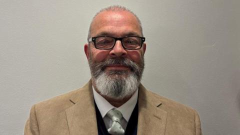A man with a grey and brown beard and glasses wearing a light brown jacket, white shirt and patterned tie.