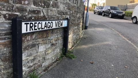 A view of Treclago View. There is a street sign and pavement, and houses in the background. There are several cars in the image.