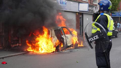 A police officer stands by a burning car during disorder in Middlesbrough on Sunday