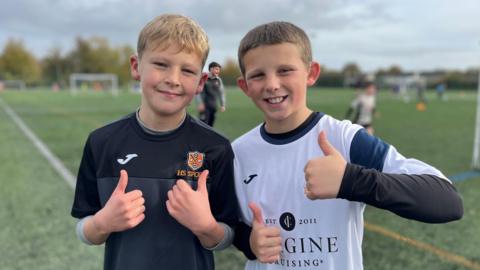 Stanley and Jacob stood next to each other on a football pitch. They are wearing football shirts and doing a thumbs up to the camera. They are both smiling and are stood by the corner flag on the football pitch. Behind them are other players on the pitch, but who are out of focus.