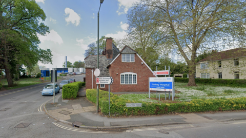 Torbay Hospital entrance pictured. The photo shows a blue welcome sign with "Torbay Hospital" written on it in white lettering. There is a small brick structure at the entrance and a road leading towards the main hospital building.