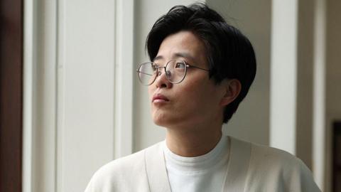 Yang Shuang-zi has short dark hair, wears thin, black rimmed glasses and wears a cream cardigan over a white t-shirt. She is staring out a window.