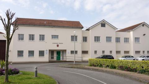 An external view of the Guernsey Prison. It is a long cream-coloured two storey building with a red roof. Every window is barred. The entrance door is green, above which is a painted shield and lettering that reads "GUERNSEY PRISON". In the foreground is a drive lined with parked cars outside the building, hedging, lawn, a pruned tree and a flower bed. 