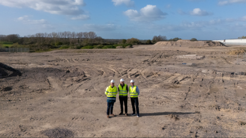 Three men in high-vis jackets and hard hats are pictured standing on a sandy building site area. The sky behind them is blue with white, fluffy clouds.