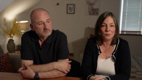 Mark Dowey in a black polo shirt leans on a table looking off to the side in thought, beside his wife Ros in a white top and black cardigan who is looking into the camera. They sit in a light lounge with a lamp on in the background and the blinds drawn.