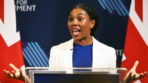 Kemi Badenoch is wearing a white jacket and a blue top. She is speaking in front of a blue background.