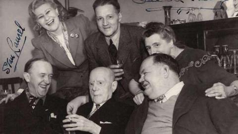 A black and white signed photo of Laurel and Hardy's visit to the Plough Inn Barkston in 1947. Three men sit in the front row of the photo, with two women and a man behind then. A cabinet full of wine glasses can be seen in the background.