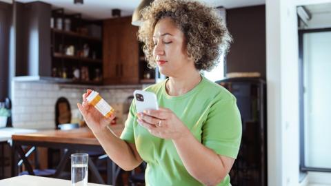 A womanholds a bottle of pills in one hand a phone in the other and looks at the pills label. She is wearing a bright green t-shirt 