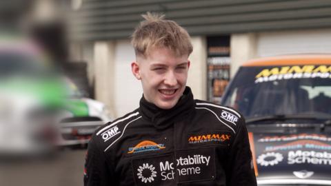 Ally Chalmers, a 17-year-old boy with short, blond hair. He is looking off to the side and is smiling. He is wearing a black race suit with the names of sponsors on it. There are racing cars behind him.