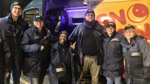 Six people, dressed in caps, and coats, standing in front of a safety van, looking at the camera and smiling. The van door is open. 