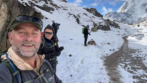 In this selfie photo, Rob Mason smiles at the camera standing in the snow somewhere on Mount Everest. He has a short grey beard and is wearing a green baseball cap with sunglasses resting on the peak. There are people standing nearby him. The ground is snowy and muddy.