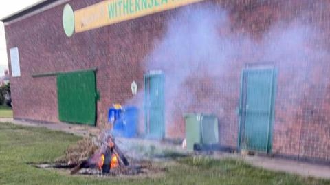 A small fire made out of wood and sticks, with smoke coming from it. It is in front of Withernsea AFC's clubhouse, which is a large building with two green, gated doors.