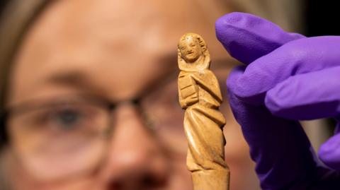 A close up of the gravoir, an ornately carved piece of red deer antler. The carving is of what looks like a hooded figure holding a book. Dr Alice Blackwell is holding the gravoir in protective purple gloves. Dr Blackwell's face is just out of focus.