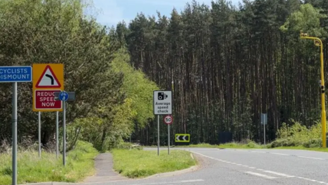 A road with a woodland to to the side of it. There are signs warning for speed cameras and a yellow tall speed camera on the right of the road.