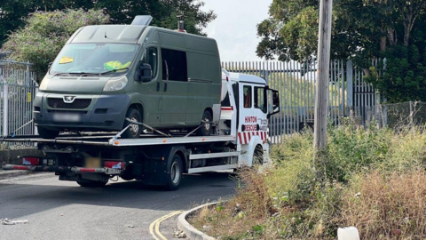 A dark green van used as a live-in vehicle is on the back of a tow truck on New Stadium Road. There is a little bit of shrubbery to the right with trees and steel fencing.