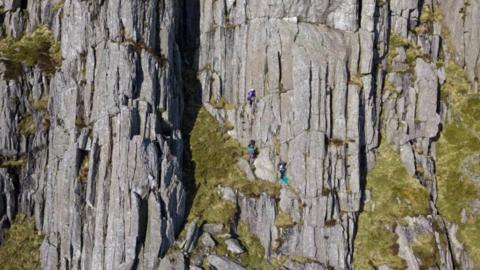 Three climbers can be seen scaling the Tryfan mountain with their safety equipment - such as harnesses and hard hats - just visible around them.