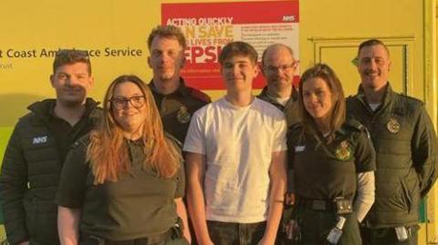 Six South East Coast Ambulance crew members standing around a teenage boy who is wearing a white shirt and grey jeans. They are standing in front of an ambulance.